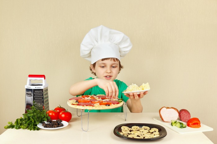 Young smiling boy in chefs hat puts a grated cheese on the pizza crust