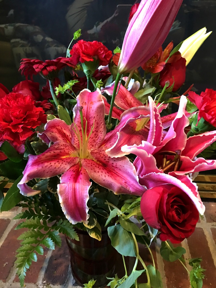 Flower arrangement in red swirl vase with Red roses, hot pink oriental lilies, red gerberas, and red carnations are arranged with pitta negra and lemon leaf flowers on a fireplace