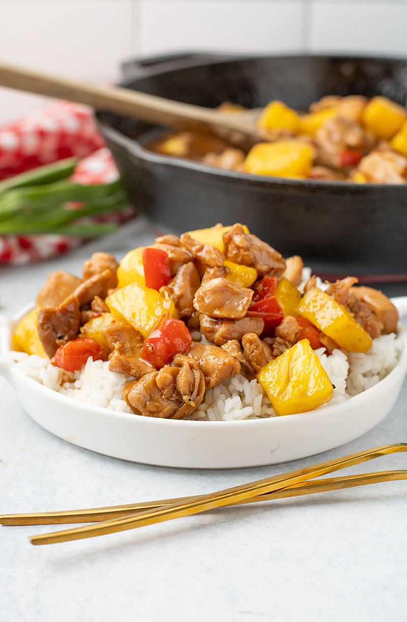 picture of freshly made pineapple chicken in a white bowl with a cast iron pan in the background