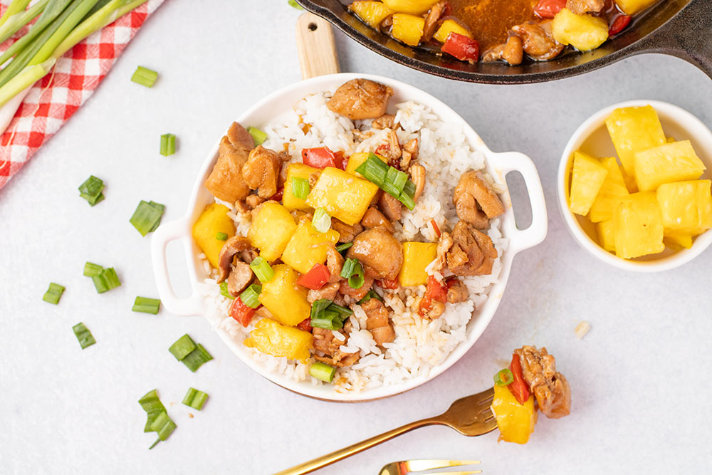 picture of freshly made pineapple chicken in a white bowl with a cast iron pan in the background
